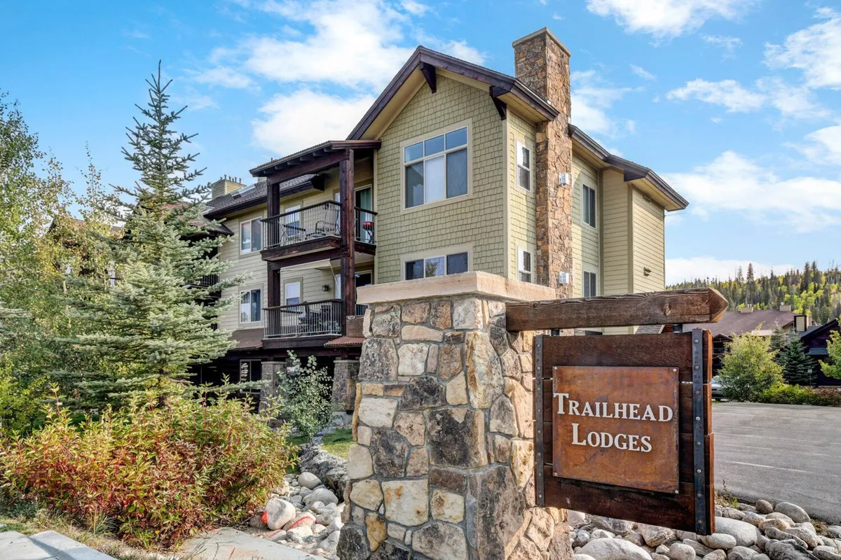 This exterior image features the stone entrance sign and wooden balconies of Trailhead Lodge Resort, set against lush green foliage. The scene radiates a warm welcome and hints at the well-maintained communal spaces where guests can relax or gather outdoors.