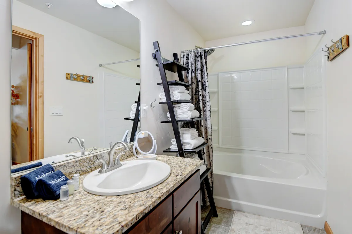 A modern bathroom with granite countertops, a large mirror, neatly stacked towels, and a spacious bathtub. The setting is bright and inviting, ideal for indulging in soothing self-care routines.