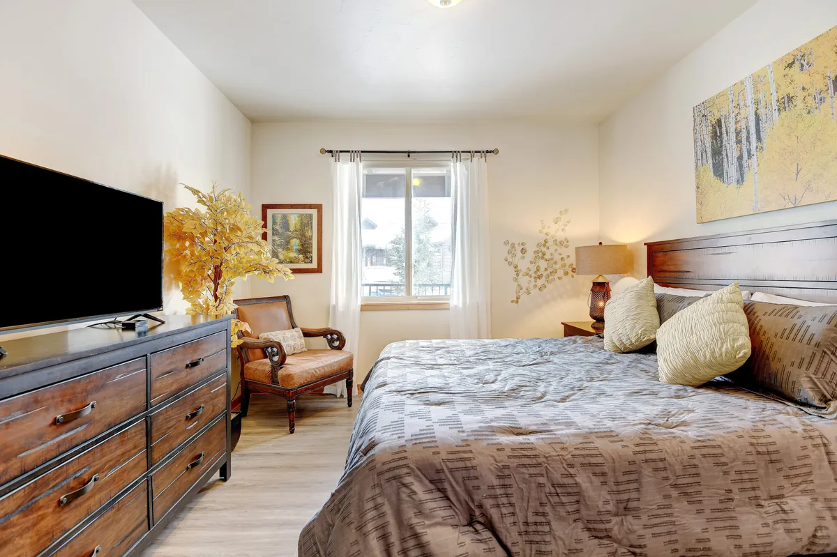 A serene bedroom featuring a large bed with decorative pillows, a sizeable dresser, an armchair set near a window draped with curtains, and calming wall art. The room exudes comfort and tranquility, ideal for relaxation after a day of adventure.