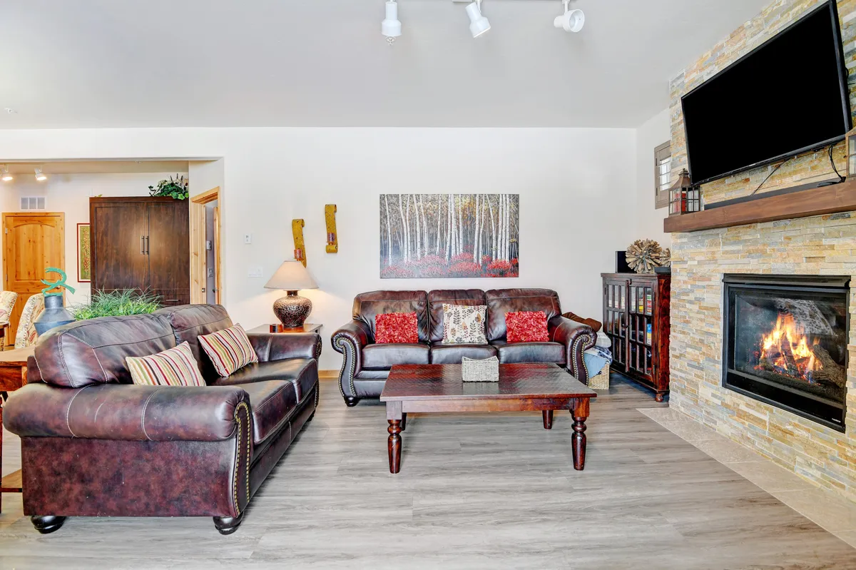 A cozy living room featuring leather sofas arranged around a wooden coffee table, with a striking fireplace as the centerpiece and artwork adorning the wall. Warm natural light fills the space, inviting guests to relax and socialize.