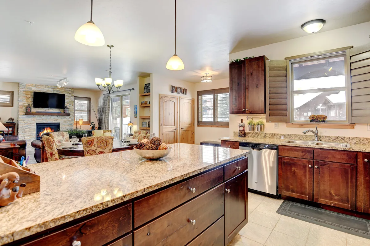 An open-concept space showcasing a granite-topped kitchen island, pendant lights, a stone-accented fireplace, wood cabinetry, and a dining area seamlessly connected to the living space. The setting is warm and sociable, ideal for meals and gatherings.
