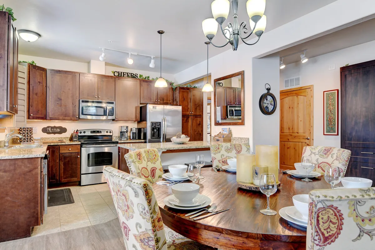 A spacious dining area with a wooden table, kitchen island, wall cabinetry, stainless steel appliances, and an elegant chandelier. The setup encourages shared meals, cooking together, and memorable family gatherings.