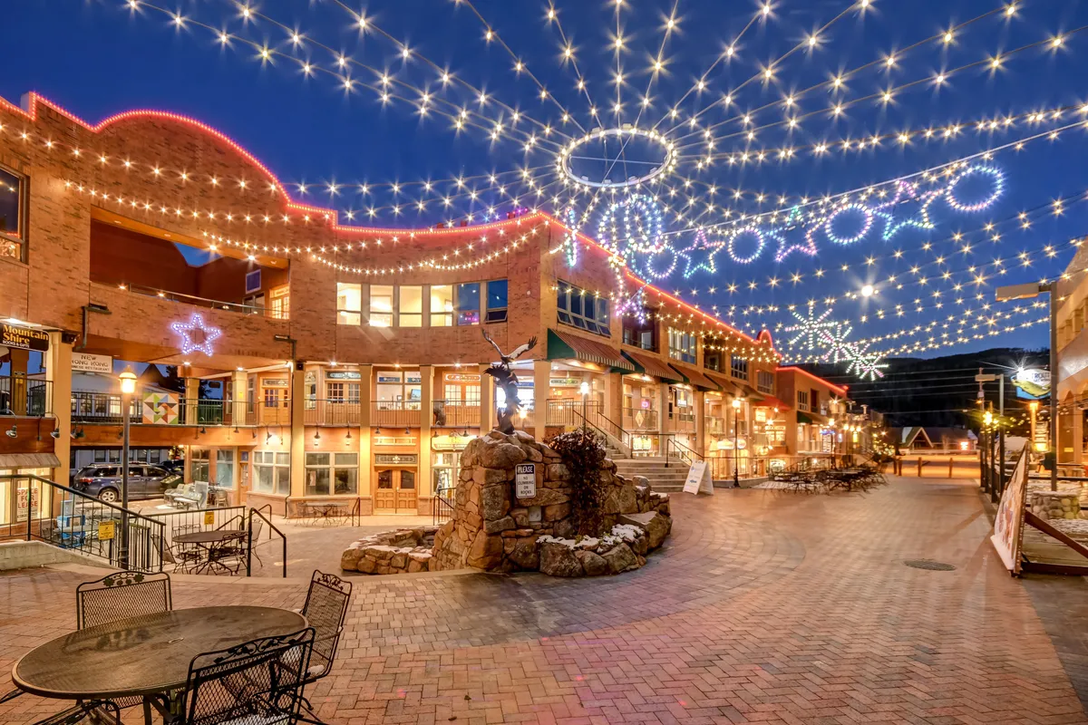 An inviting exterior shot featuring twinkling lights hung above a stone fountain and brick pathway, surrounded by decorative stars. The atmosphere is festive and vibrant, suggesting a community space ideal for evening strolls or socializing with family and pets.