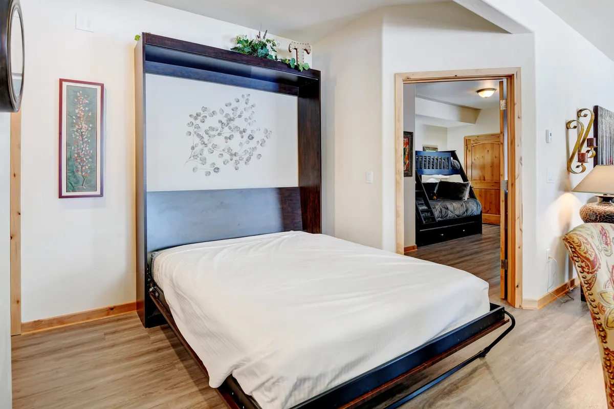 A versatile living space featuring a Murphy bed cleverly integrated into the room, decorative artwork, warm wood accents, and an open doorway leading to another room. The setup maximizes flexibility without sacrificing style.