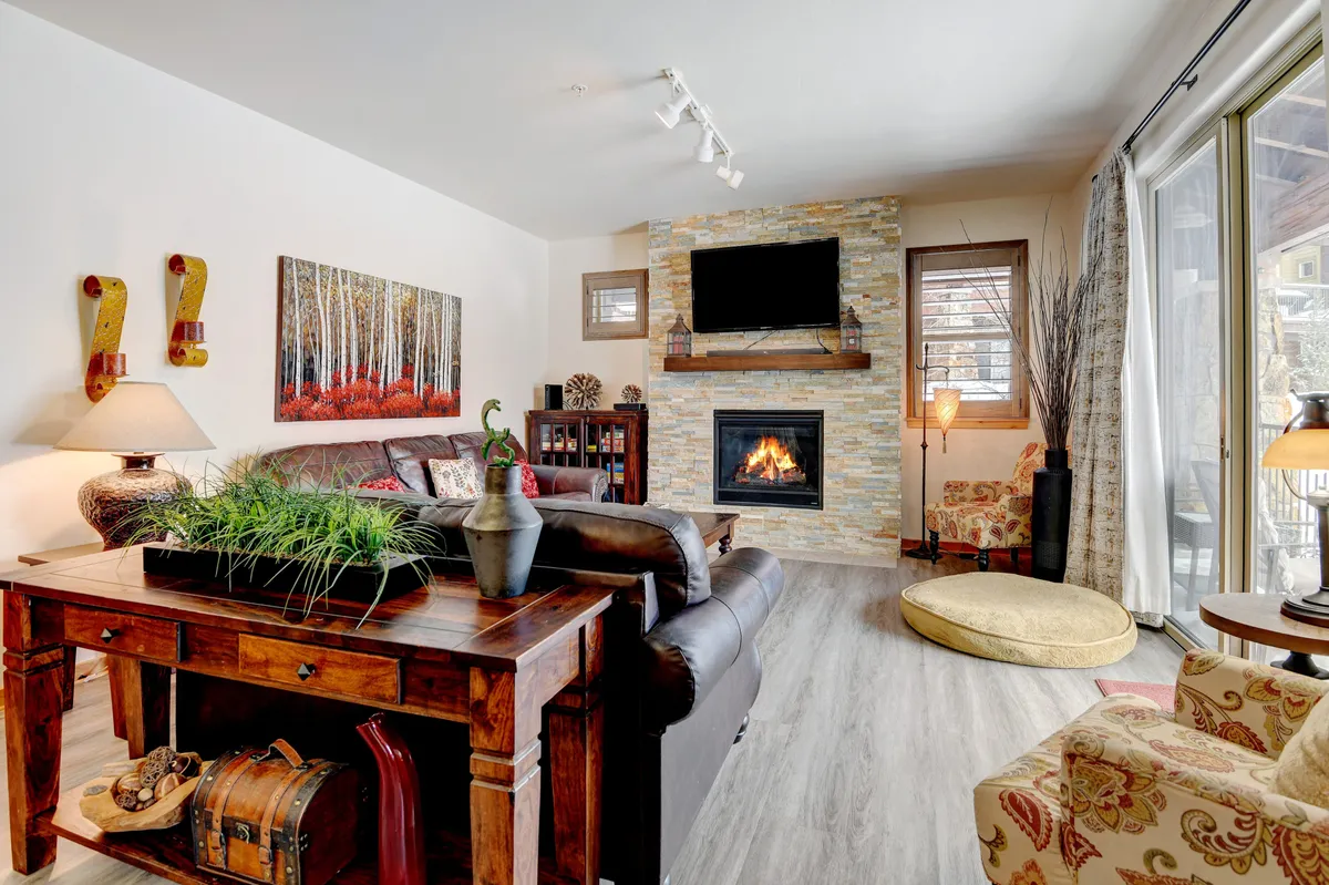 The image showcases a cozy and inviting living area inside a luxury condo, featuring a glowing fireplace as the focal point. Comfortable seating surrounds a wooden table, and modern artwork adds an elegant touch to the space. The scene radiates warmth and relaxation, perfect for unwinding after a day of mountain adventures.